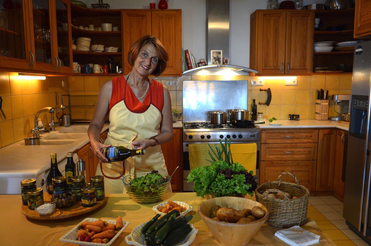 Küche, auf der Anrichte stehen Schalen mit Karotten, Zucchini, Kartoffel und grünem Salat sowie ein Tablett mit verschiedenen Produkten der Olive. Daninter steht Burgi Bläuel und gießt Olivenöl in den Salat. 