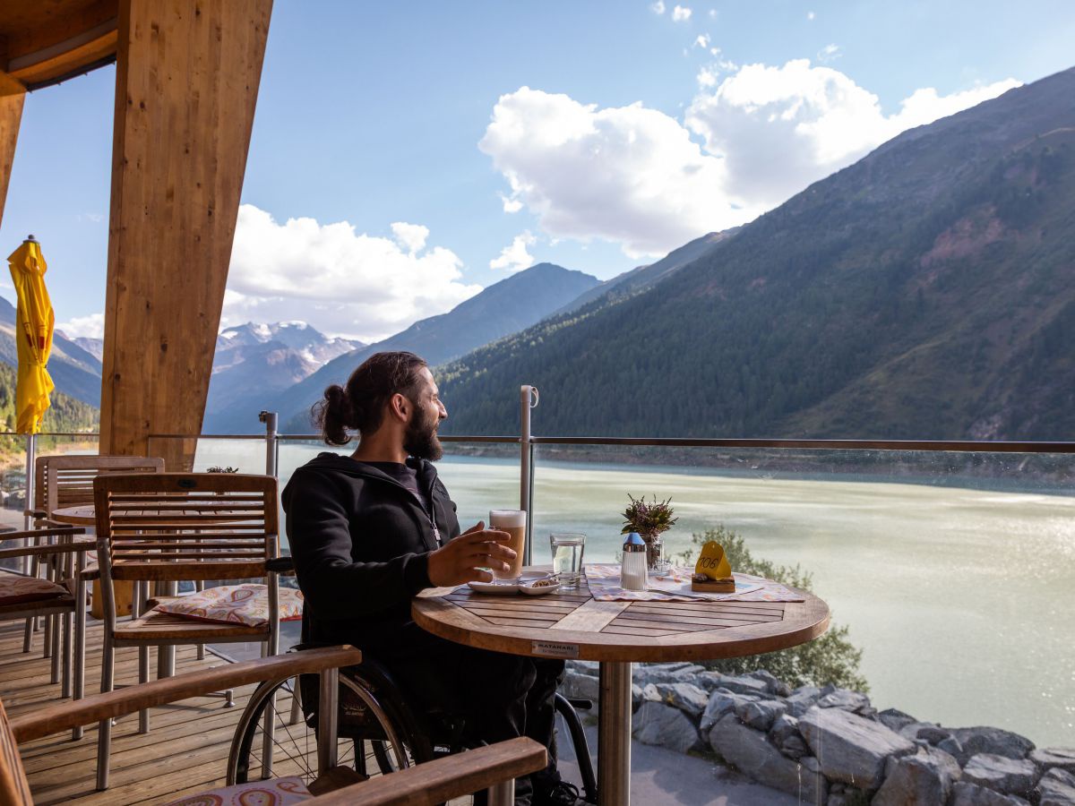 Ein Mann mit langem zusammengebundenem Haar und Bart genießt auf einer Terrasse den Ausblick über einen Gebirgssee. Er sitzt in einem Rollstuhl an einem runden Holztischchen und trinkt Kaffee aus einem Glas.